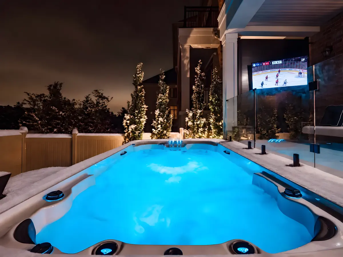 Illuminated swim spa on a snowy backyard deck with an outdoor TV and glass railing at night