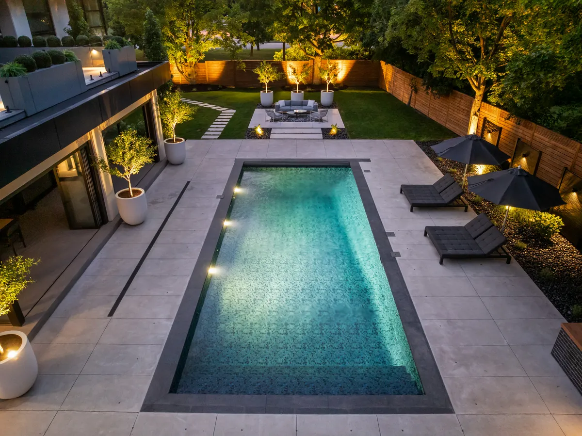 Modern backyard pool with porcelain patio, lounge seating, planters and landscape lighting at dusk