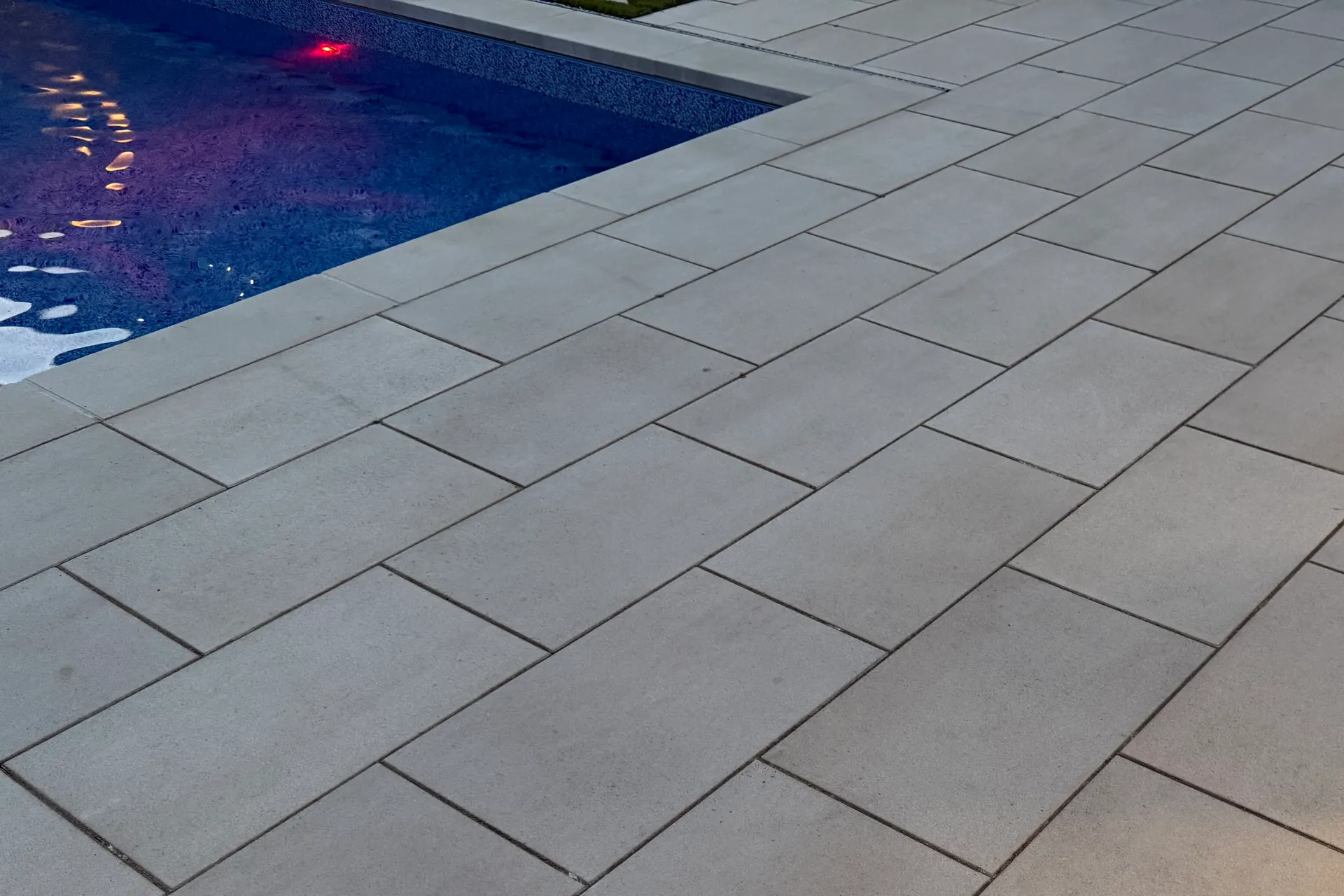 Backyard pool surrounded by a large-format paver patio with landscape lighting at dusk