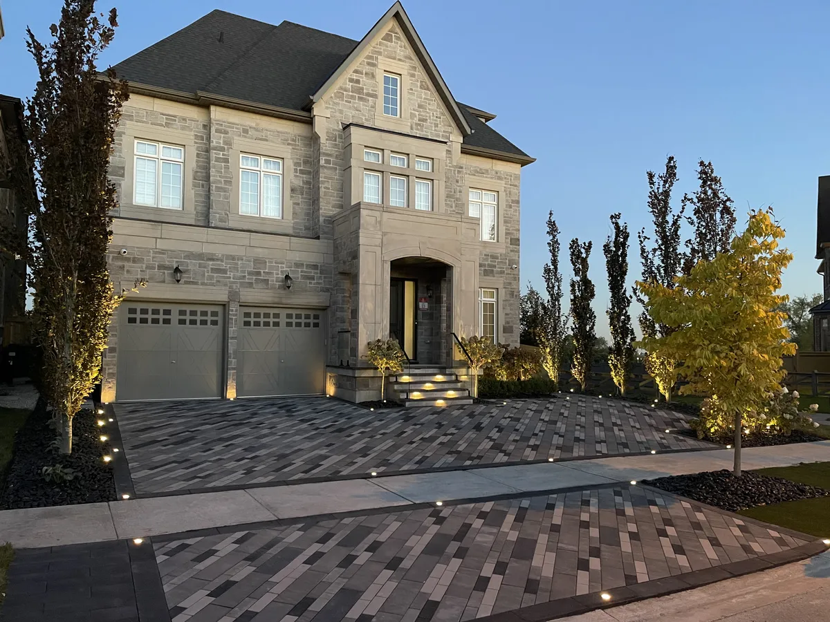 Interlocking paver driveway with a mixed-tone linear pattern and in-ground lighting in front of a stone home