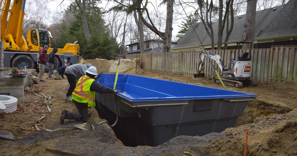Crew guiding a fibreglass pool shell into an excavated backyard during crane delivery