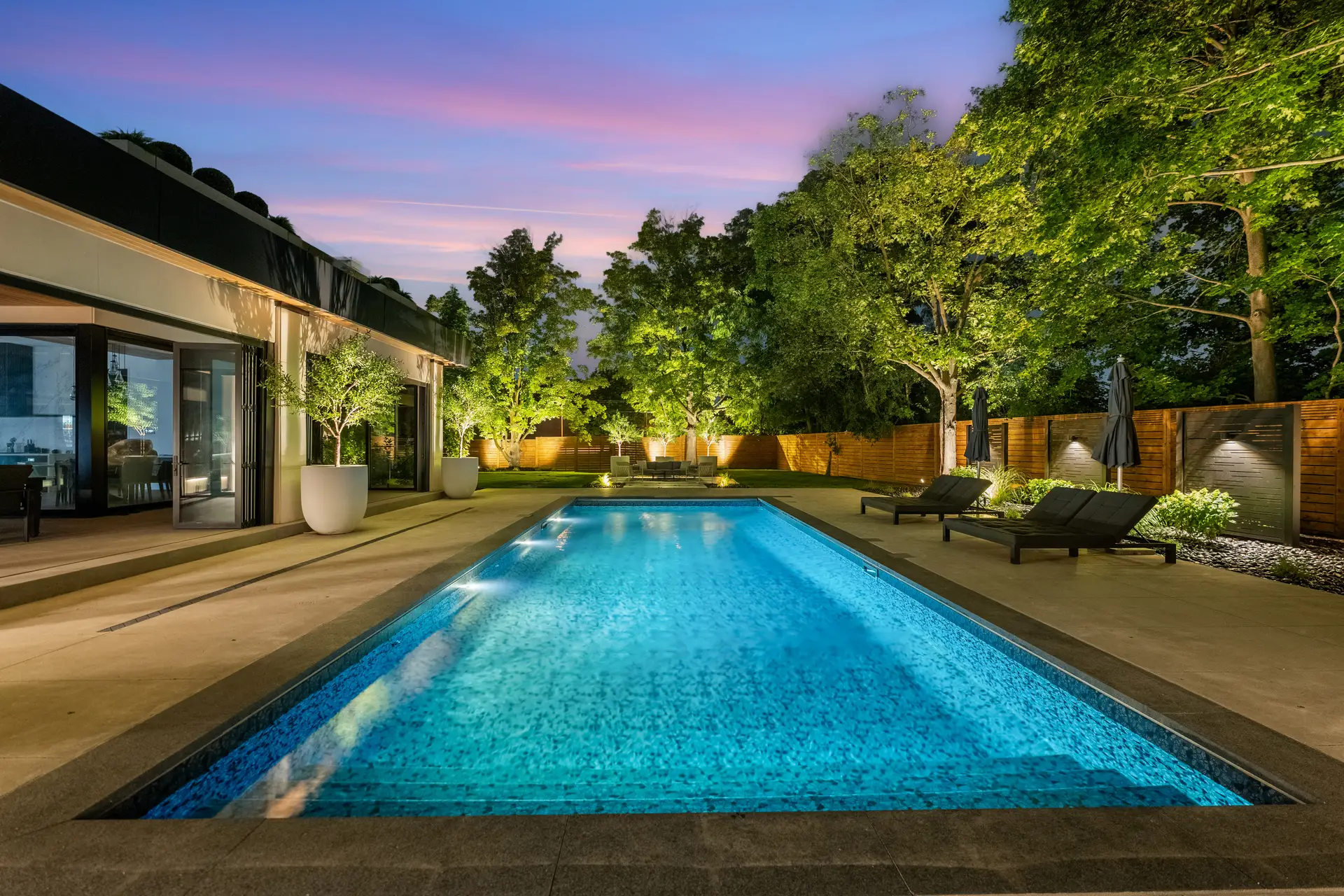 Backyard pool at dusk with lounge seating, landscape lighting, and cedar privacy fencing behind a modern Toronto home