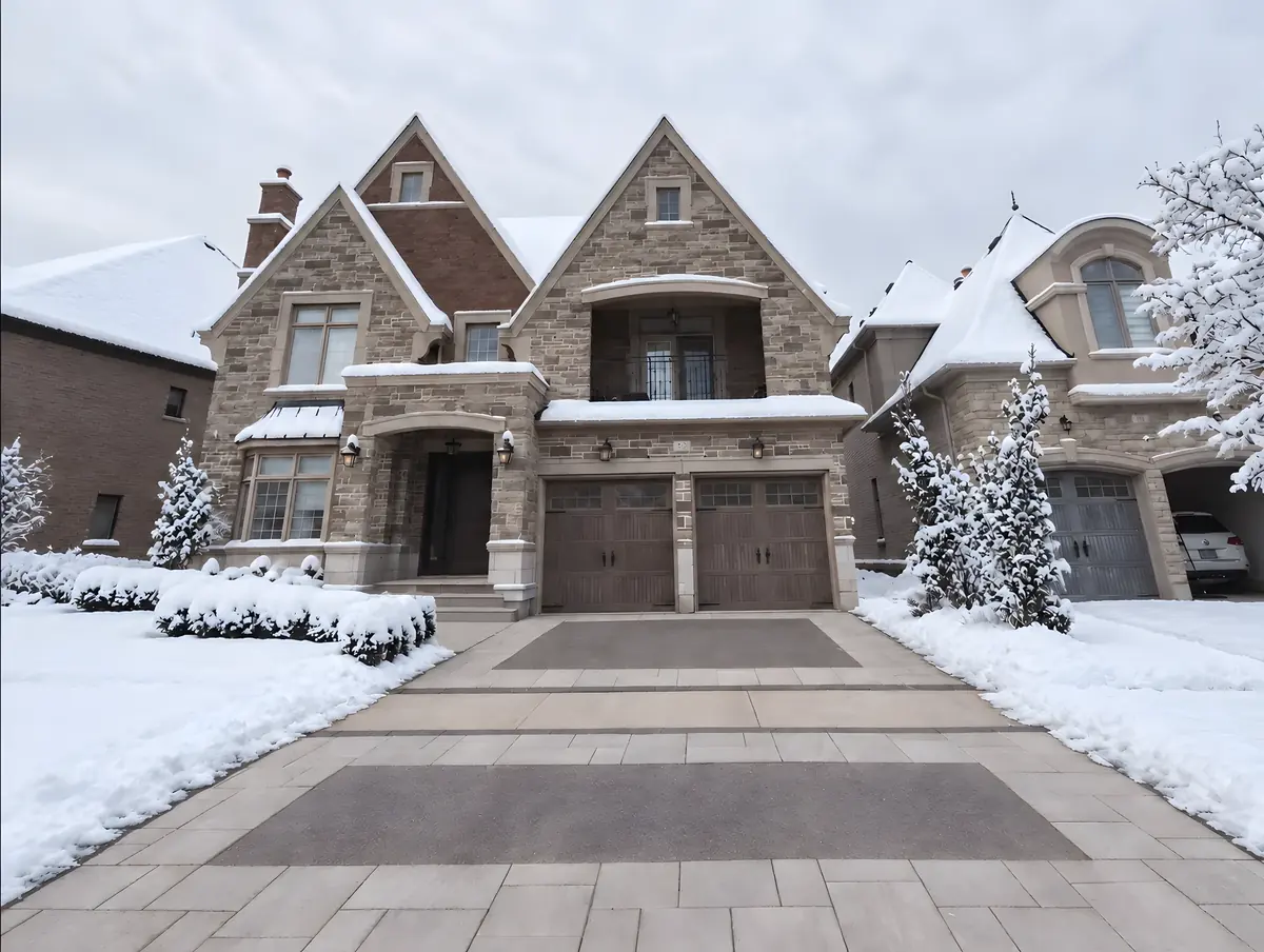 Clear two-tone paver driveway in front of a stone home with snow-covered lawns and evergreens on either side