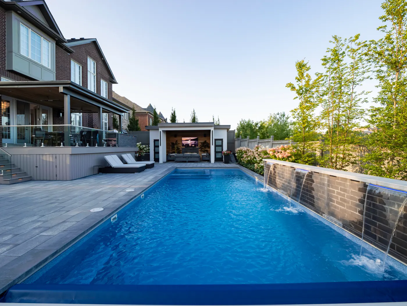 Rectangular fiberglass pool with sheer descent water features along a stone wall and a cabana beyond